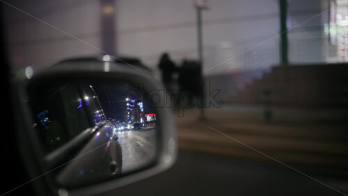 View in a car side mirror reflecting the road behind at night, headlights and streetlights stretching into the distance