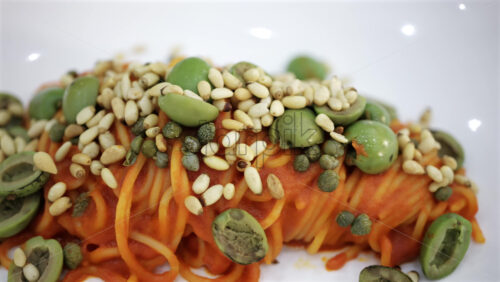 Close up of a pasta dish with tomato sauce topped with green olives, seeds, and capers, presented in a modern gourmet style