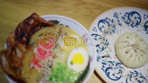 Ramen bowl served with sliced pork, soft boiled egg, noodles, greens, and sesame seeds next to a steamed bun