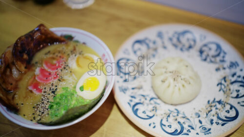 Ramen bowl served with sliced pork, soft boiled egg, noodles, greens, and sesame seeds next to a steamed bun