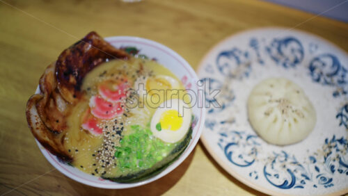 Ramen bowl served with sliced pork, soft boiled egg, noodles, greens, and sesame seeds next to a steamed bun