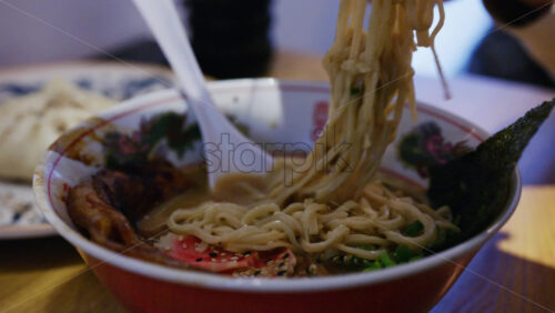 Hand lifting noodles from a ramen bowl with chopsticks while seated at a table