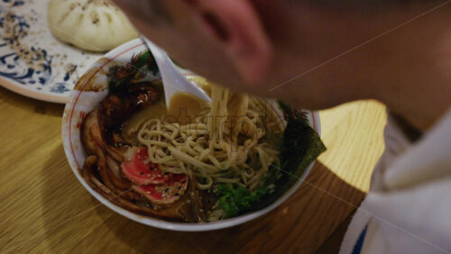 Hand lifting noodles from a ramen bowl with chopsticks while seated at a table
