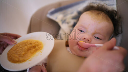 Infant seated in a high chair while an adult feeds the baby with a spoon