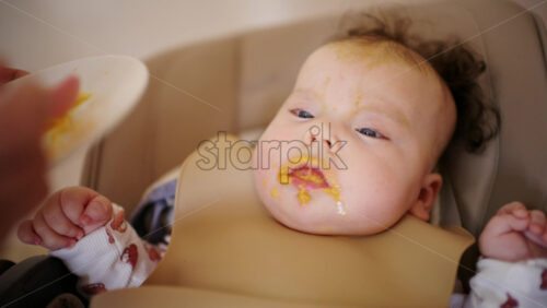 Infant seated in a high chair while an adult feeds the baby with a spoon