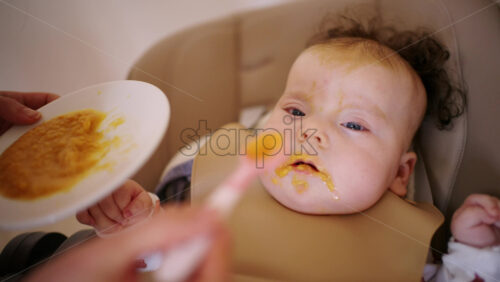 Infant seated in a high chair while an adult feeds the baby with a spoon