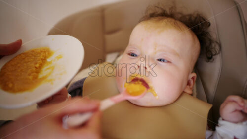 Infant seated in a high chair while an adult feeds the baby with a spoon