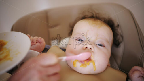 Infant seated in a high chair while an adult feeds the baby with a spoon