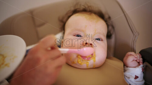Infant seated in a high chair while an adult feeds the baby with a spoon