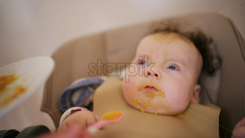 Infant seated in a high chair while an adult feeds the baby with a spoon