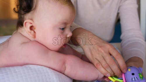 Mother playing with baby using a colorful rattle while the infant lies on a soft cushion