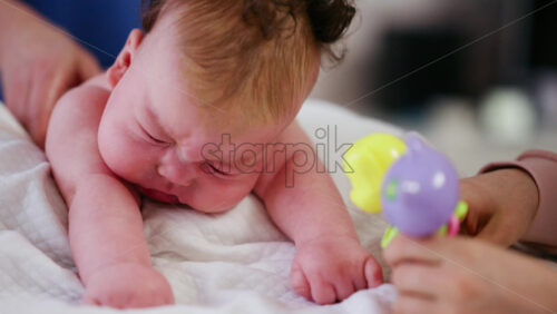 Woman trying to distract a crying baby lying on a blanket during massage with a toy