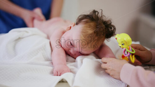 Woman trying to distract a crying baby lying on a blanket during massage with a toy