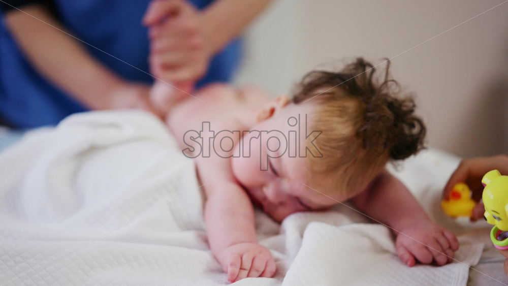 Woman trying to distract a crying baby lying on a blanket during massage with a toy