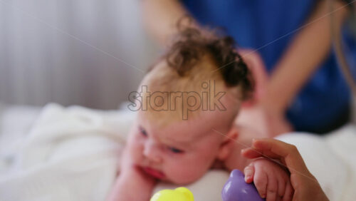 Woman trying to distract a crying baby lying on a blanket during massage with a toy