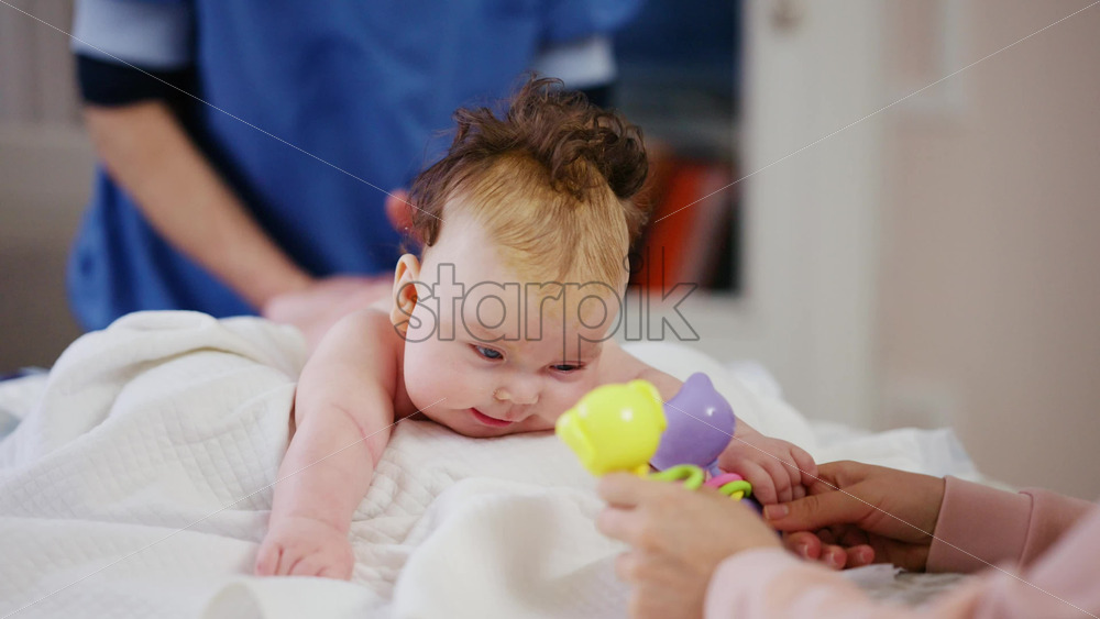 Woman trying to distract a crying baby lying on a blanket during massage with a toy