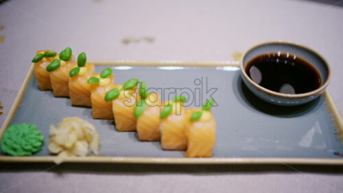 A plated dish of salmon sushi, neatly arranged with green garnish on top with soy sauce in a small bowl, plus wasabi and pickled ginger on the side