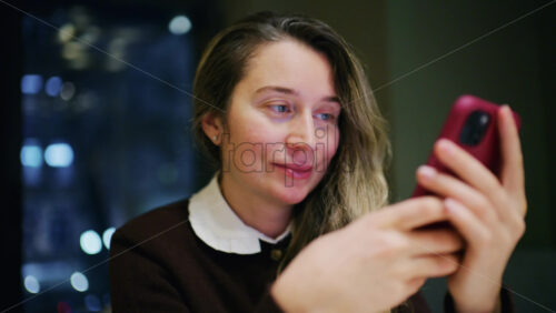 A woman indoors at night, softly lit, looking at her phone with a slight smile