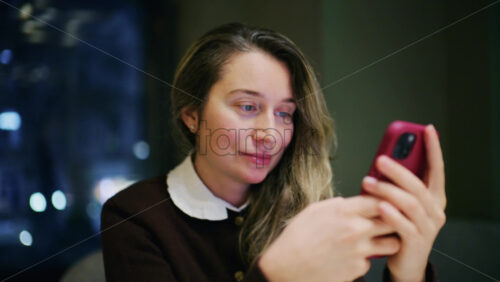 A woman indoors at night, softly lit, looking at her phone with a slight smile