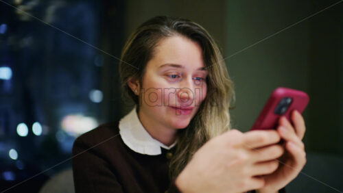 A woman indoors at night, softly lit, looking at her phone with a slight smile