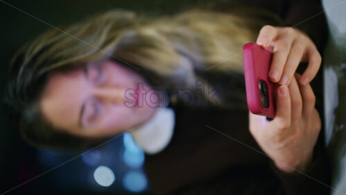 A woman indoors at night, softly lit, looking at her phone with a slight smile. Vertical