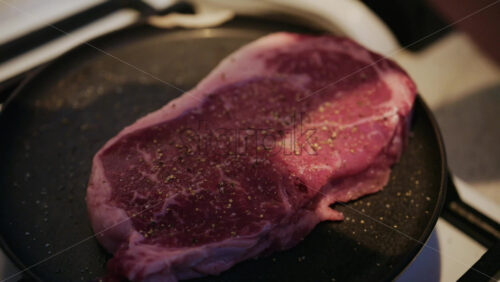 Close up of a raw beef steak being seasoned with salt and black pepper in a pan