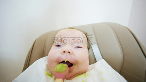Close up of a baby eating green vegetable puree from a spoon while sitting in a high chair