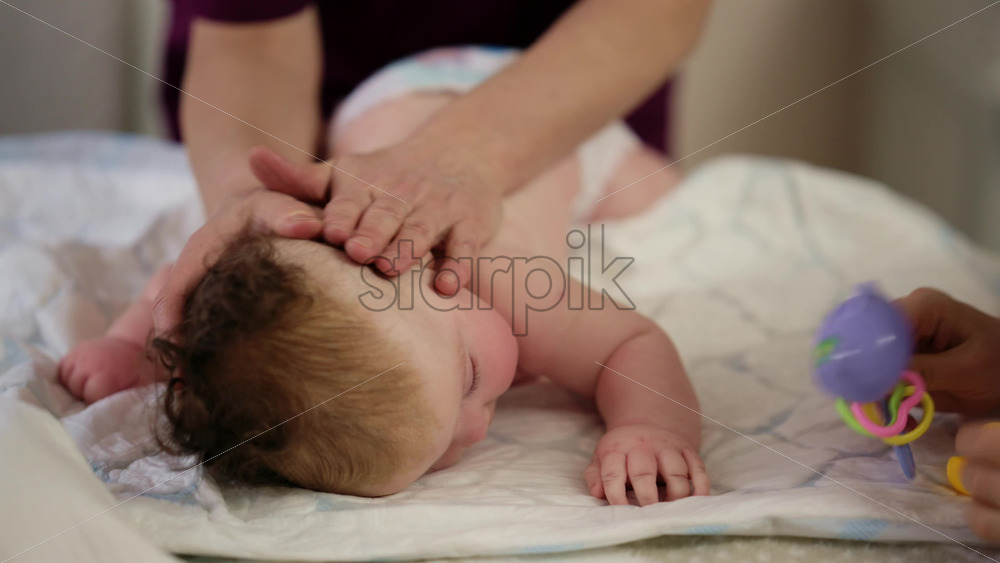 Woman trying to distract a crying baby lying on a blanket during massage with a toy
