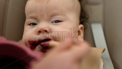 Infant seated in a high chair while an adult feeds the baby with a spoon