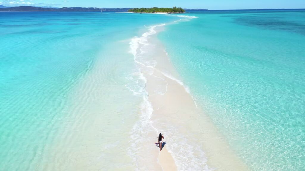Video – Aerial drone view of a woman walking along a narrow white sandbar between shallow turquoise waters, with a small island visible in the distance in Madagascar - Starpik Stock
