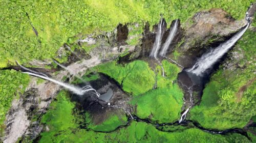Video – Aerial drone view of multiple waterfalls cascading down a lush green cliff into a rocky basin, surrounded by dense tropical rainforest vegetation in Madagascar - Starpik Stock