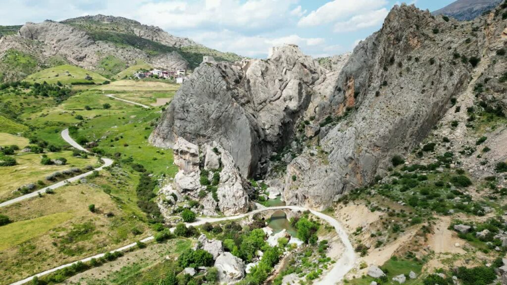 Video – Aerial drone view of rocky mountains and canyon landscapes with a small stone bridge crossing a river below, surrounded by rugged terrain and green valley vegetation - Starpik Stock