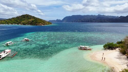 Video – Aerial drone view of traditional Filipino bangka boats anchored near a white sand beach in Coron, Palawan, Philippines. Crystal clear shallow waters transition from light aqua to deep blue with limestone islands in the distance - Starpik Stock