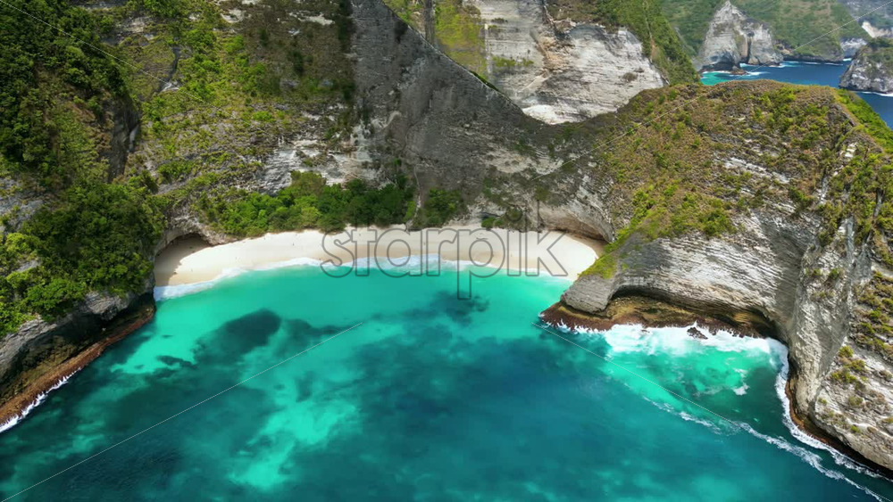 Video - Aerial drone view of Kelingking Beach in Nusa Penida, Bali, Indonesia. Dramatic limestone cliffs tower above a pristine white sand beach and vibrant turquoise water. The famous T-Rex shaped headland dominates the tropical coastline under bright daylight