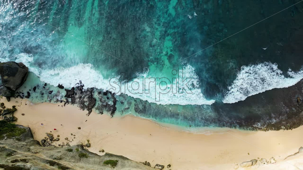 Video - Aerial drone view of Diamond Beach in Nusa Penida, Bali. Crystal clear ocean waves break against dramatic cliff walls and white sand beach below. Limestone formations and vibrant blue water create a stunning tropical island landscape