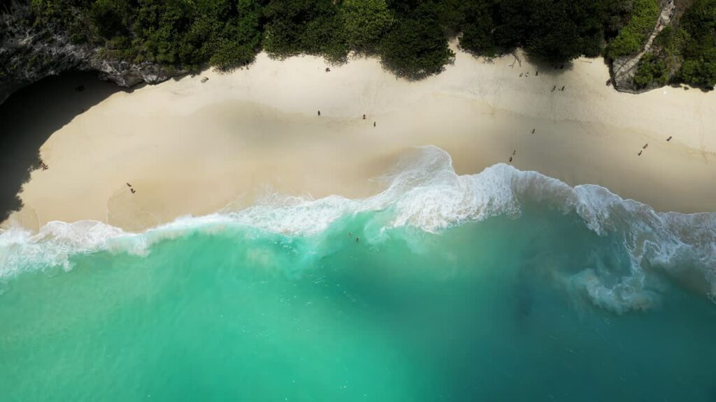 Video - Aerial drone view of Kelingking Beach in Nusa Penida, Bali. White sand shoreline meets crystal clear turquoise ocean waves rolling onto the beach