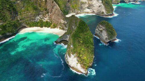 Video - Aerial drone view of Kelingking Beach in Nusa Penida, Bali, Indonesia. Dramatic limestone cliffs tower above a pristine white sand beach and vibrant turquoise water. The famous T-Rex shaped headland dominates the tropical coastline under bright daylight