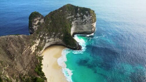Video - Aerial drone view of the iconic T-Rex cliff formation at Kelingking Beach, Nusa Penida, Bali. Steep limestone cliffs drop into the deep blue Indian Ocean, with waves crashing against the secluded beach below