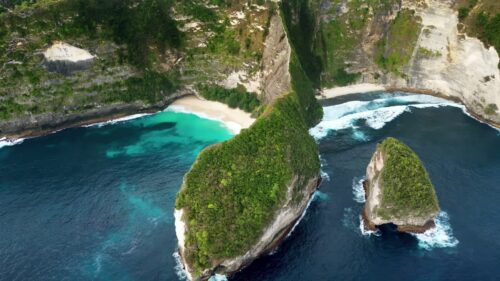 Video - Aerial drone view of Kelingking Beach in Nusa Penida, Bali, Indonesia. Dramatic limestone cliffs tower above a pristine white sand beach and vibrant turquoise water. The famous T-Rex shaped headland dominates the tropical coastline under bright daylight