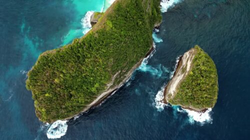 Video - Aerial drone view of Kelingking Beach in Nusa Penida, Bali, Indonesia. Dramatic limestone cliffs tower above a pristine white sand beach and vibrant turquoise water. The famous T-Rex shaped headland dominates the tropical coastline under bright daylight