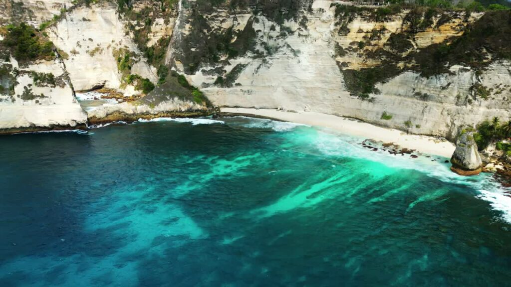 Video - Aerial drone view of Atuh Beach in Nusa Penida, Bali, Indonesia. Turquoise water reveals coral reef patterns beneath the surface, surrounded by high limestone cliffs and a quiet white sand shoreline