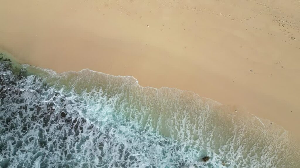 Video - Aerial drone view of waves crashing onto Diamond Beach, Nusa Penida, Bali. White foamy surf rolls over turquoise water and smooth sand shoreline
