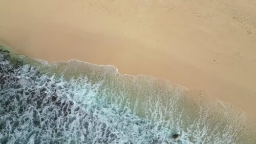 Video - Aerial drone view of waves crashing onto Diamond Beach, Nusa Penida, Bali. White foamy surf rolls over turquoise water and smooth sand shoreline