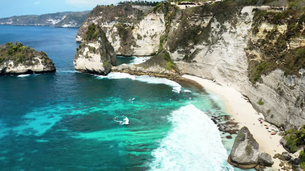 Video - Aerial drone view of Diamond Beach and its iconic offshore rock pinnacle in Nusa Penida, Bali. Powerful ocean waves meet dramatic limestone cliffs and lush tropical vegetation