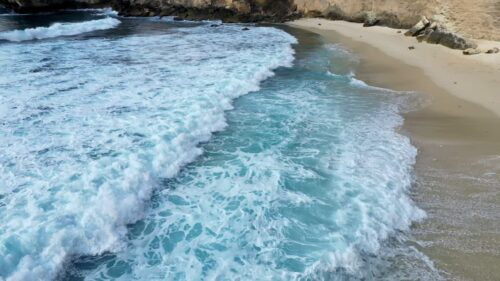 Video - Aerial drone view of ocean waves gently washing onto a sandy beach in Nusa Penida, Bali, Indonesia. Foamy surf creates natural patterns over turquoise water and light sand