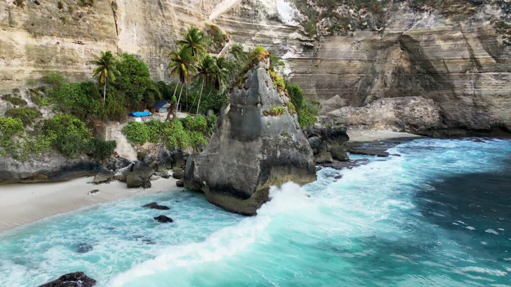 Video - Aerial drone view of the iconic limestone rock formation at Diamond Beach, Nusa Penida, Bali, Indonesia. Turquoise waves crash against the massive sea stack surrounded by towering white cliffs and tropical palm trees