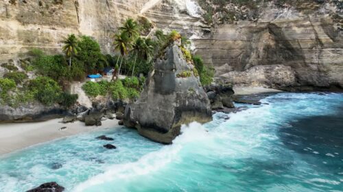 Video - Aerial drone view of the iconic limestone rock formation at Diamond Beach, Nusa Penida, Bali, Indonesia. Turquoise waves crash against the massive sea stack surrounded by towering white cliffs and tropical palm trees