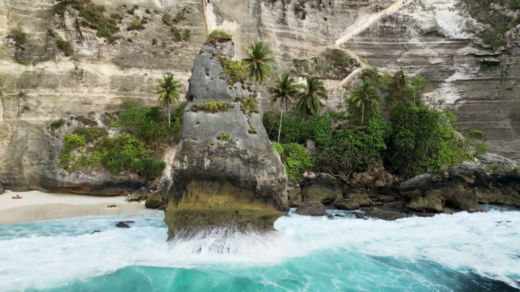 Video - Aerial drone view of the iconic limestone rock formation at Diamond Beach, Nusa Penida, Bali, Indonesia. Turquoise waves crash against the massive sea stack surrounded by towering white cliffs and tropical palm trees