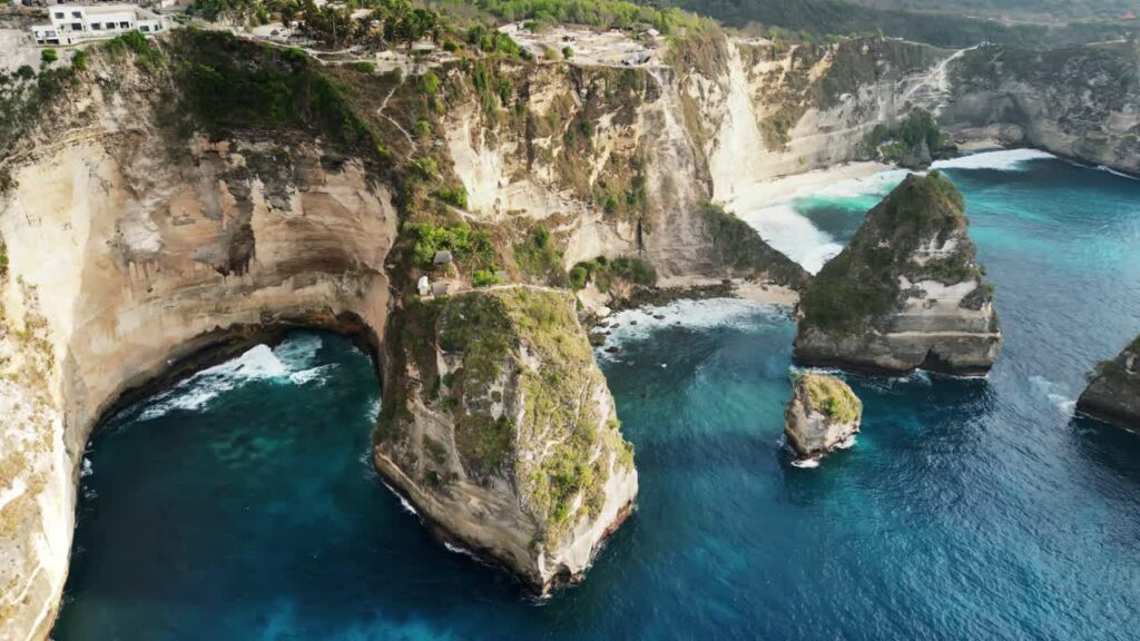 Video - Aerial drone view of Kelingking Beach in Nusa Penida, Bali, Indonesia. Dramatic limestone cliffs tower above a pristine white sand beach and vibrant turquoise water. The famous T-Rex shaped headland dominates the tropical coastline under bright daylight