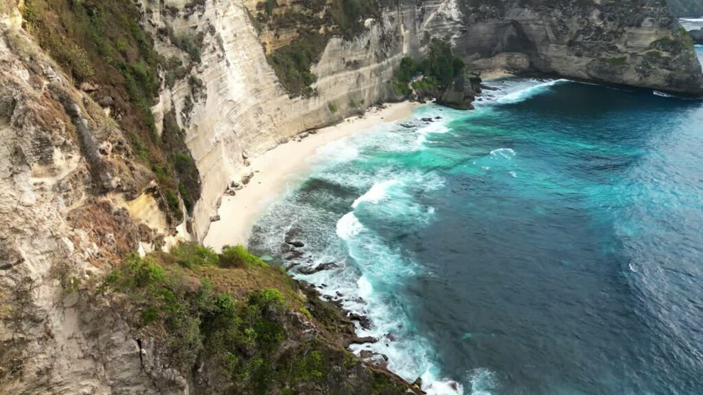 Video - Aerial drone view of Diamond Beach in Nusa Penida, Bali. Secluded white sand shoreline curves beneath steep limestone cliffs as turquoise ocean waves roll in from the Indian Ocean